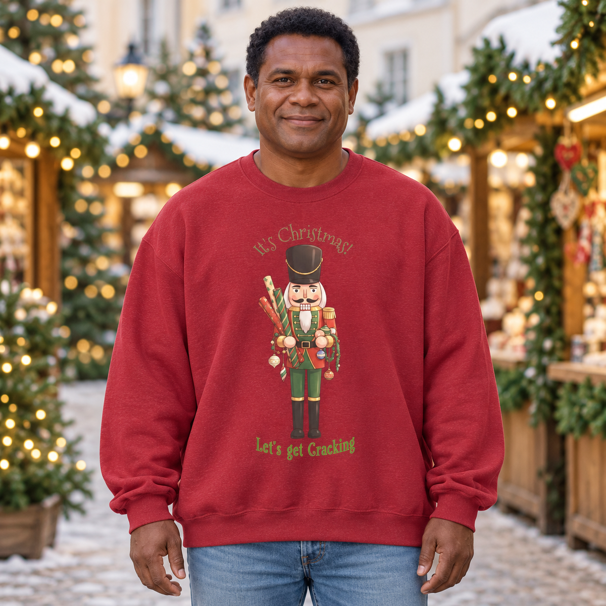 A man wearing a red crewneck sweatshirt with a Christmas nutcracker graphic and the text 'It's Christmas! Let's get Cracking' stands in front of a festive outdoor holiday market background with lights and trees.