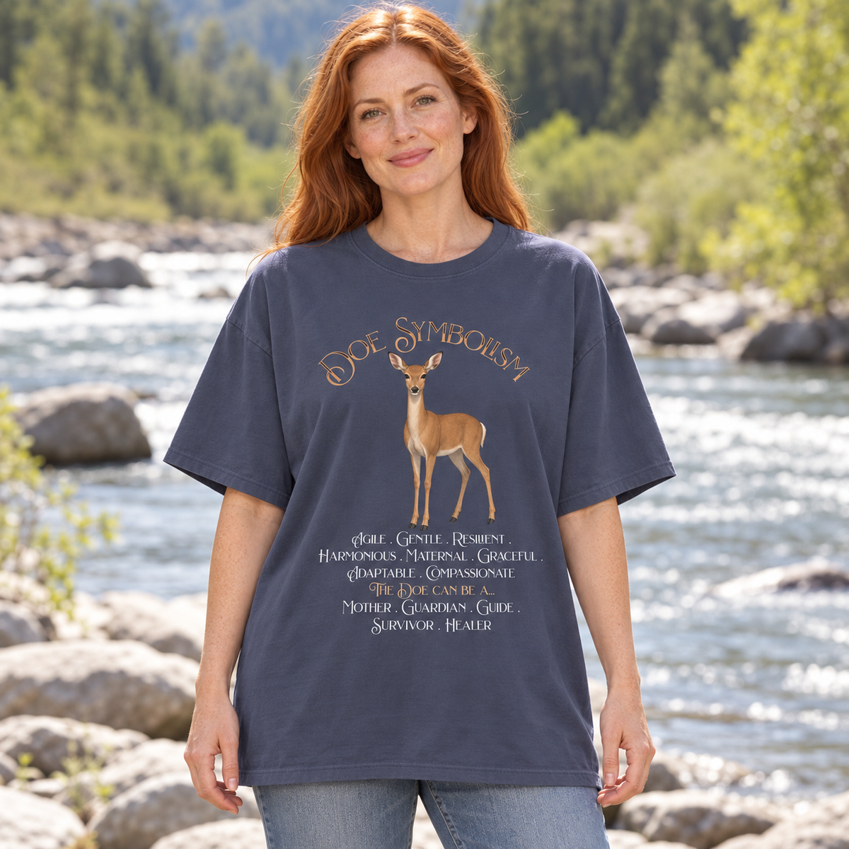 A woman standing outdoors by a river wearing a navy graphic T-shirt with a deer illustration and the text 'Doe Symbolism' and descriptive script.
