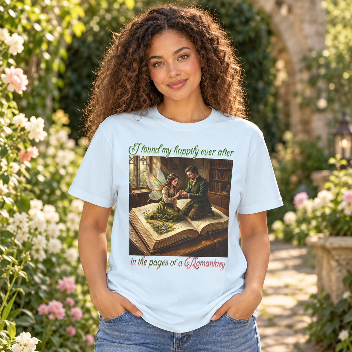 A woman with curly brown hair wearing a light blue graphic t-shirt and blue jeans, standing in a garden with flowers and a stone path in the background. The t-shirt features a central illustration of a man putting a ring on a fairy's finger as they sit on an open book, with the text 'I found my happily ever after in the pages of a Romantasy'.