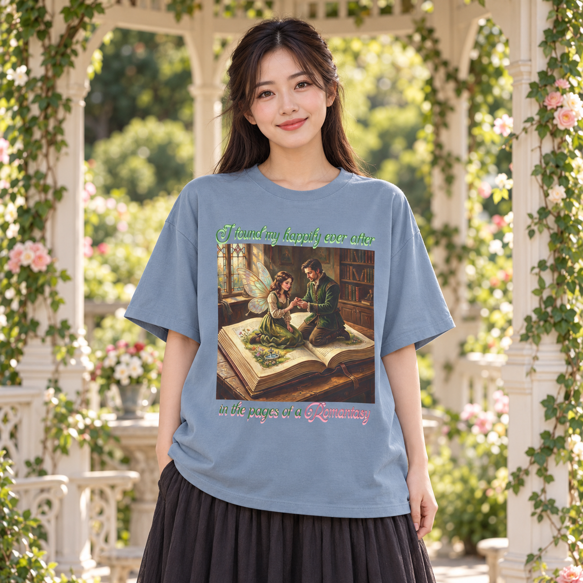 A woman wearing a light blue graphic t-shirt with a vintage-style illustration of a man proposing to a fairy as they sit on an open book in a library. The woman is posed in front of a white gazebo with green foliage and roses.