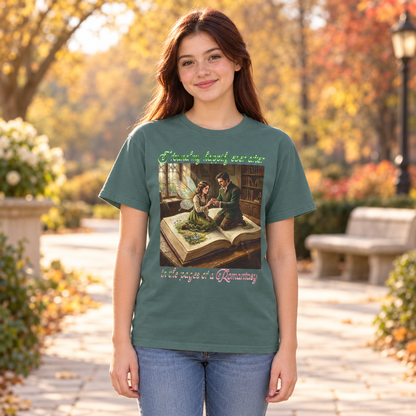 A young woman stands outdoors on a sunlit campus walkway wearing a muted green graphic T-shirt and blue jeans. The shirt features a vintage-style illustration of a man proposing to a fairy as they sit on a large book in a library, with decorative text above and below the artwork that says, I found my happily ever after in the pages of a Romantasy.