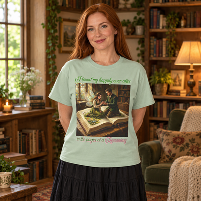 A woman with red hair stands in a cozy, book-filled library wearing a light sage green graphic t-shirt. The t-shirt features a vintage-style illustration of a man proposing to a fairy as they sit on a book and the text 'I found my happily ever after in the pages of a Romantasy'.
