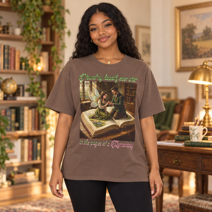 A woman stands in a cozy, book-filled room wearing a brown graphic t-shirt. The t-shirt features a vintage-style illustration of a man proposing to a fairy as they sit on an open book, with green and pink text reading 'I found my happily ever after in the pages of a Romantasy'.