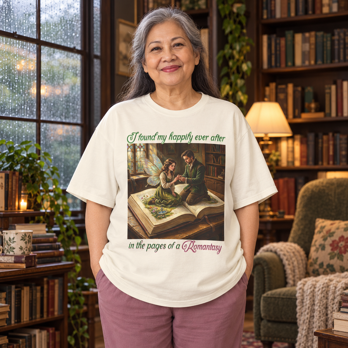 An older woman stands in a cozy home library wearing a cream-colored graphic t-shirt. The t-shirt features a central illustration of a man proposing to a fairy as they sit on an open book, with the text 'I found my happily ever after in the pages of a Romantasy' printed around the graphic.