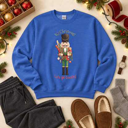 A royal blue crewneck sweatshirt with a nutcracker holding wrapping paper and ornaments surrounded by the words, 'It's Christmas! Let's go Christmas!' text. The pullover is styled in a flat lay with a box of holiday decorations, gray sweatpants, a wristwatch, brown slippers, and gray socks on a light beige background.