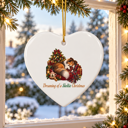 A heart-shaped white ceramic Christmas ornament hanging from a gold string, featuring an illustration of a sleeping Sheltie by a decorated Christmas tree with the text 'Dreaming of a Sheltie Christmas'. The ornament is shown hanging in front of a snowy window with Christmas lights and pine branches.