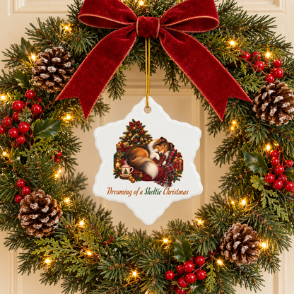 A festive Christmas wreath with a large red velvet bow, green pine branches, warm white string lights, red berries, and pinecones. A white snowflake-shaped ornament hangs from the center, featuring an illustration of a sleeping sheltie clutching a book under a Christmas tree with the text 'Dreaming of a Sheltie Christmas'.