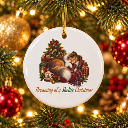 A round white ceramic Christmas ornament hanging on a decorated tree. The ornament features an illustration of a sleeping sable sheltie beside a small Christmas tree, a steaming mug filled with whip, and gift boxes, with the text 'Dreaming of a Sheltie Christmas' below.