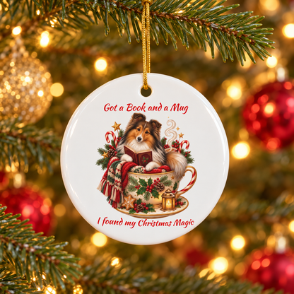 A round white Christmas ornament hanging from a gold string on a decorated tree. The ornament features an illustration of a fluffy dog reading a book in a mug, with red text reading 'Got a Book and a Mug' and 'I found my Christmas Magic'.