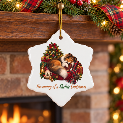 A white snowflake-shaped Christmas ornament hanging from a gold string in front of a fireplace mantel decorated with pine branches, red plaid ribbon, and warm lights. The ornament features an illustration of a sleeping sheltie on a red bed and plaid blanket beside a decorated tree and gift boxes, with the text 'Dreaming of a Sheltie Christmas' below.