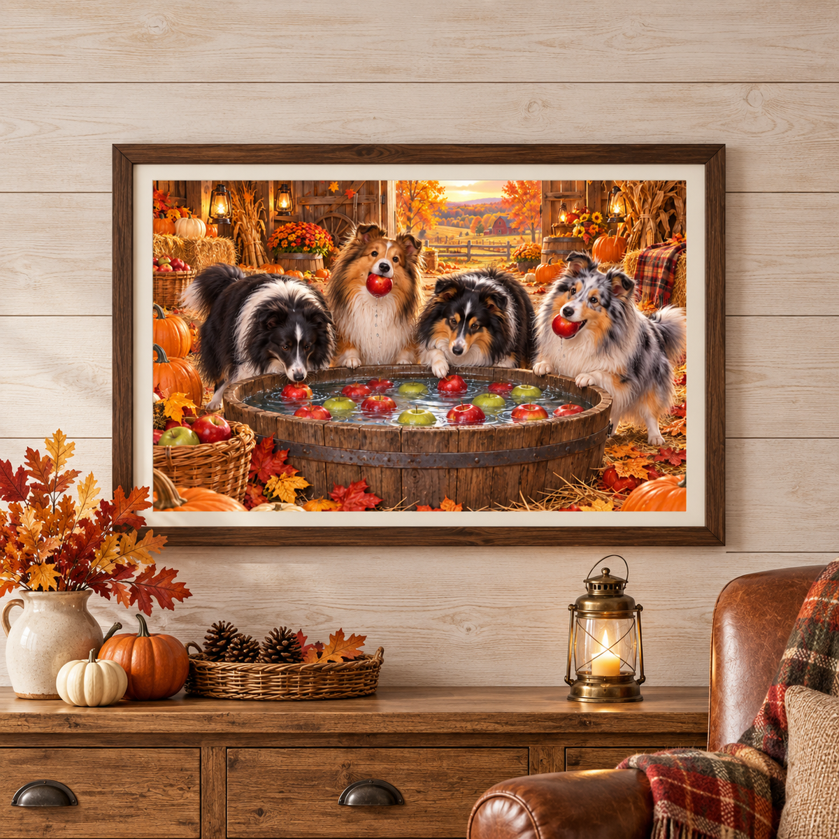 A framed wall art print showing four shelties bobbing for apples, displayed in a cozy autumn-themed barn on a white wood wall above a wooden console table with pumpkins, a lantern, a basket of pinecones, and a leather armchair.