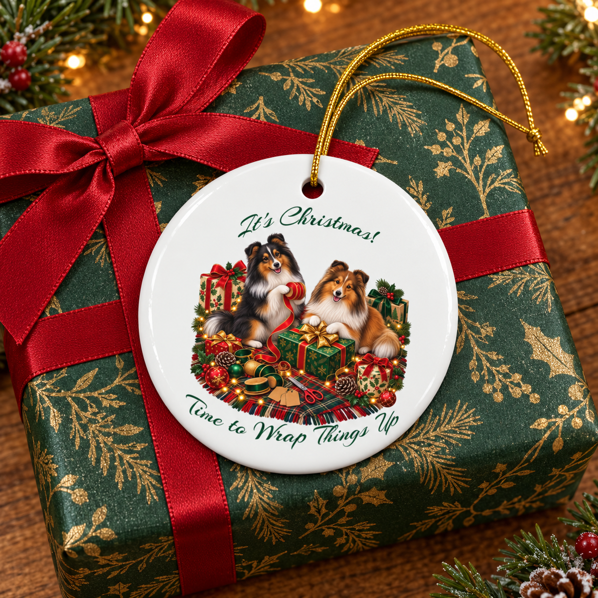 A round white Christmas ornament with a gold hanging string, featuring an illustration of two shelties surrounded by holiday gifts and greenery, with the text 'It's Christmas! Time to Wrap Things Up'. The ornament is displayed on top of a wrapped gift box with a red ribbon and green and gold wrapping paper.