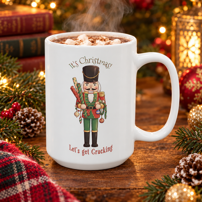 A white ceramic mug filled with a hot cocoa sits on a wooden table surrounded by Christmas decorations, including pine branches, ornaments, books, and a plaid blanket. The mug features a nutcracker illustration with the text 'It's Christmas! Let's get Cracking'.