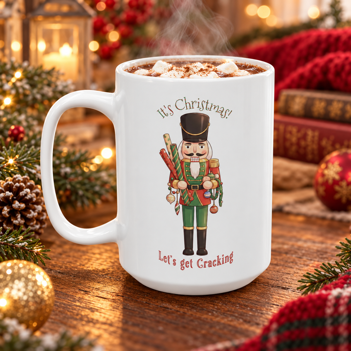 A white ceramic mug filled with hot cocoa sits on a wooden table surrounded by Christmas decorations, including pine branches, ornaments, books, and a red knit blanket. The mug features a printed nutcracker illustration with the text 'It's Christmas! Let's get Cracking'.