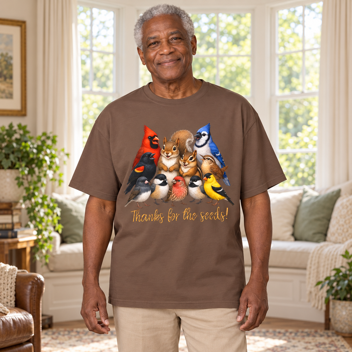 An older man wearing a brown graphic t-shirt with cartoon birds, a chipmunk, and a squirrel and the text 'Thanks for the seeds!' The man stands in a bright living room.