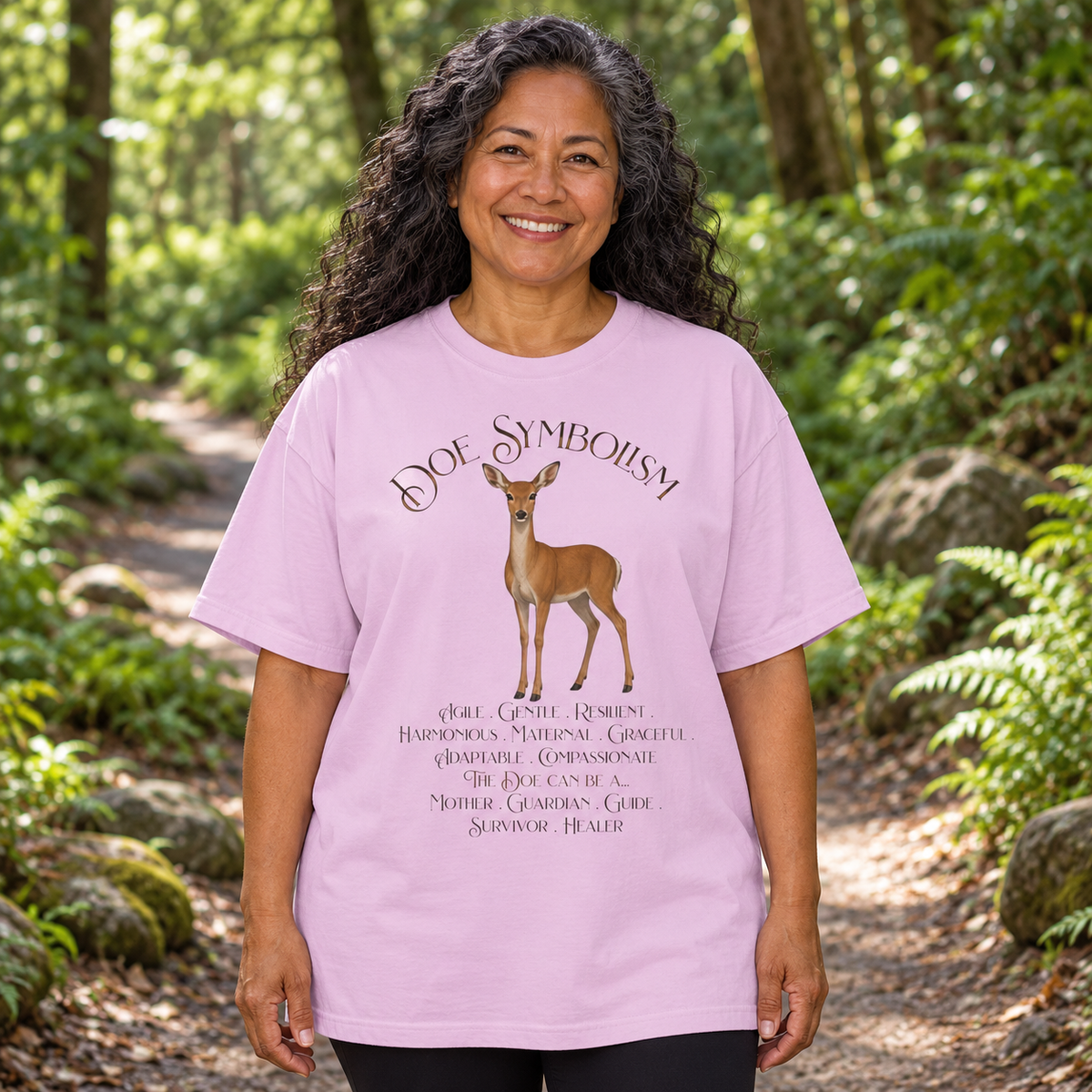 A woman standing on a forest path wearing a light purple graphic t-shirt with a deer illustration and text that says, 'Doe Symbolism'.