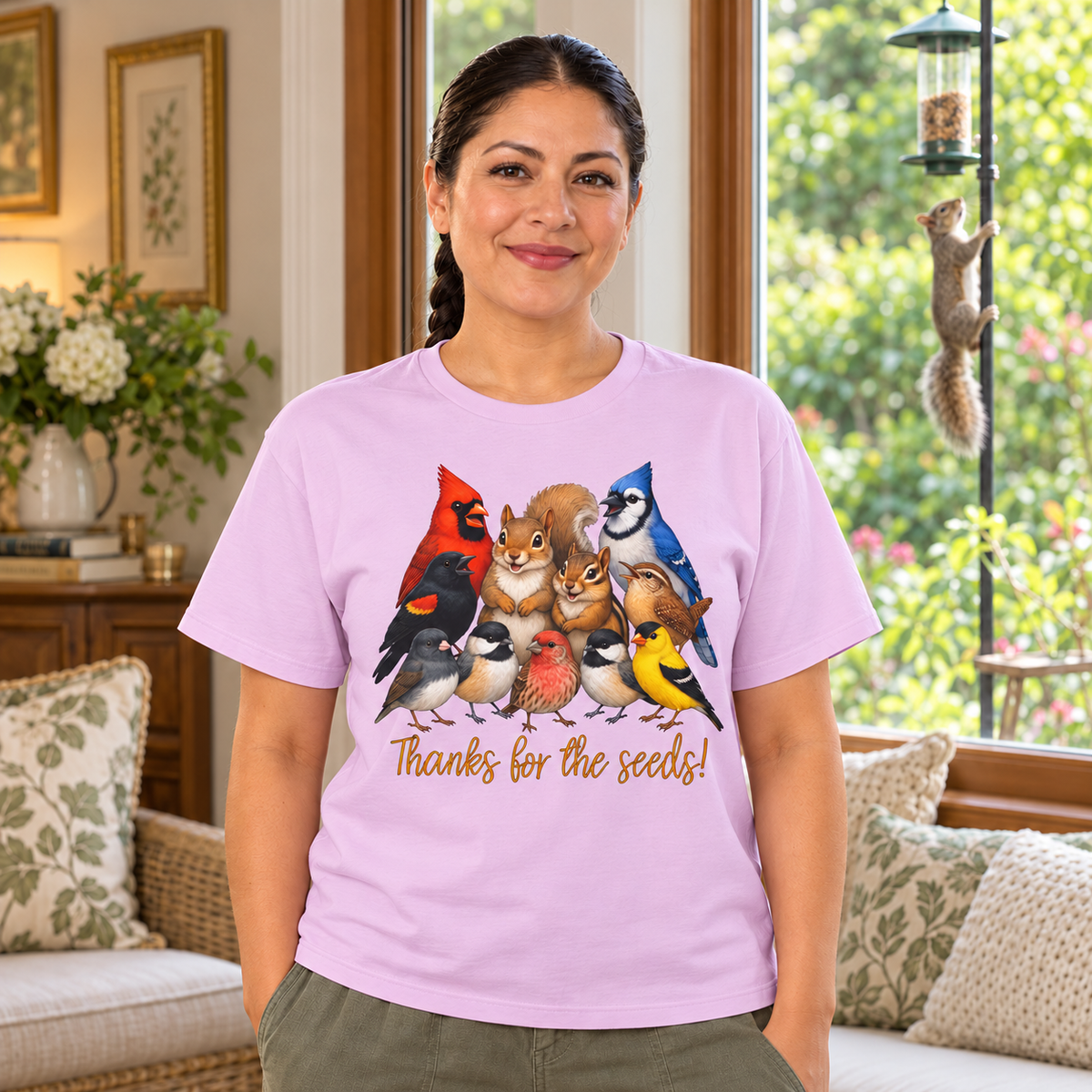 A woman wearing a light purple graphic t-shirt with a colorful illustration of various birds, chipmunk, and a squirrel standing as if in a group shot, with the text 'Thanks for the seeds!' printed below. She is standing in a cozy, sunlit living room with a couch, pillows, and a window looking out onto green foliage.
