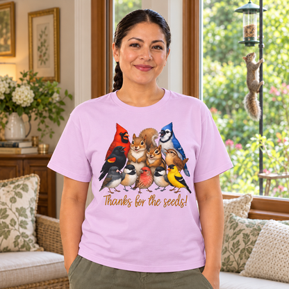 A woman wearing a light purple graphic t-shirt with a colorful illustration of various birds, chipmunk, and a squirrel standing as if in a group shot, with the text 'Thanks for the seeds!' printed below. She is standing in a cozy, sunlit living room with a couch, pillows, and a window looking out onto green foliage.