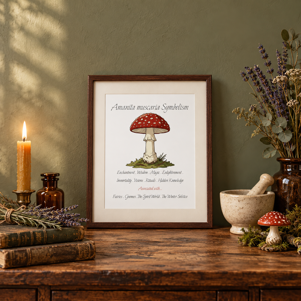 A framed art print featuring a red and white spotted mushroom with text reading 'Amanita muscaria Symbolism' is displayed on a rustic wooden table. The scene is styled with a lit candle, books, dried lavender, mortar and pestle, vase of dried flowers, and a small matching mushroom figurine against a textured olive green wall.