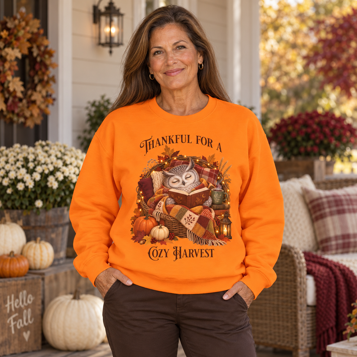 A woman stands on a fall decorated front porch wearing a bright orange crewneck sweatshirt with a graphic of an owl reading a book surrounded by pumpkins, autumn leaves, and the text 'Thankful For A Cozy Harvest'.