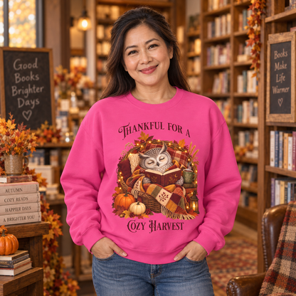 A woman wearing a bright pink sweatshirt with an autumn owl graphic and the text 'Thankful For A Cozy Harvest'. She stands in a cozy bookshop setting.