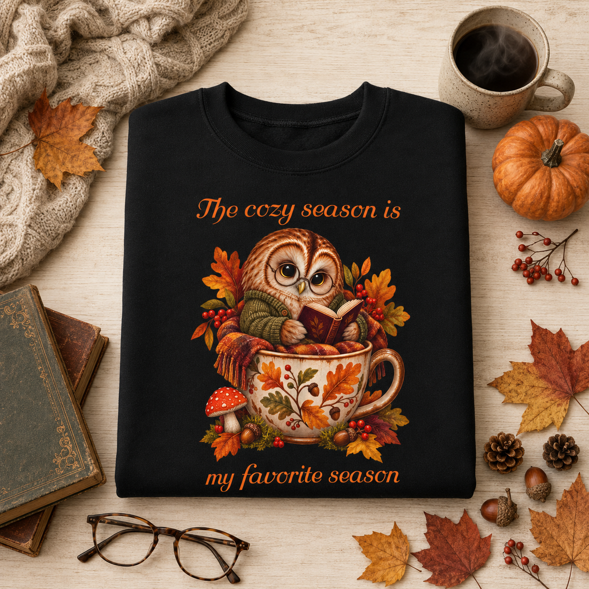 A black crewneck sweatshirt folded on a light wood surface, featuring an autumn-themed owl illustration reading 'The cozy season is my favorite season.' The sweatshirt is styled with fall props including leaves, a pumpkin, books, glasses, a scarf, and a mug of coffee.