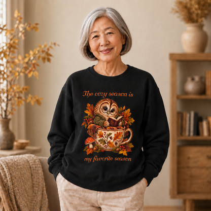 An older woman stands indoors wearing a black crewneck sweatshirt with a colorful autumn owl graphic and the text, 'The cozy season is my favorite season.' The room has warm neutral tones, a wooden bookshelf, and dried plants in the background.