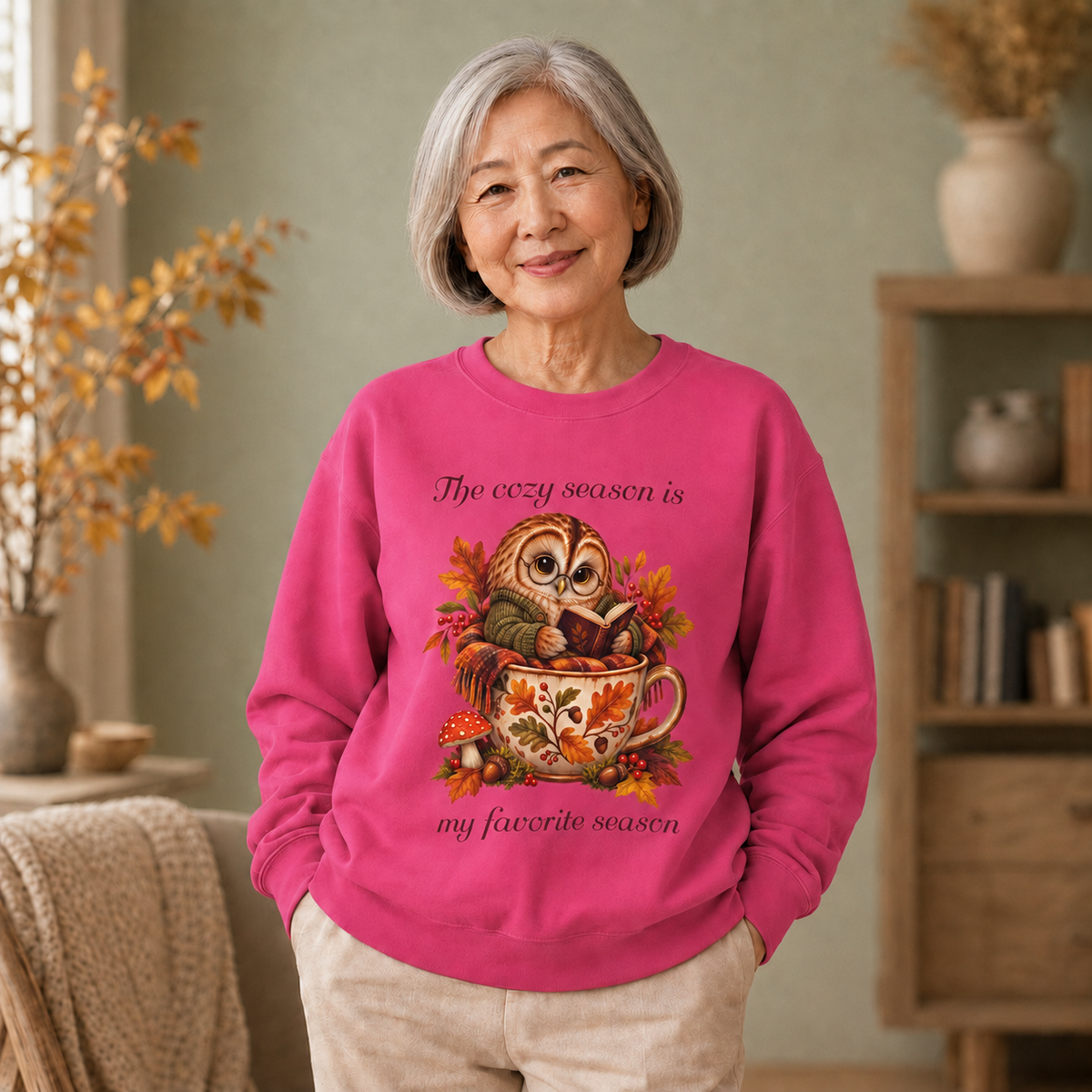 An older woman stands indoors wearing a bright pink crewneck sweatshirt with a large autumn owl graphic and the text, 'The cozy season is my favorite season.' The room has a soft neutral background with shelves, a vase, and a blanket.