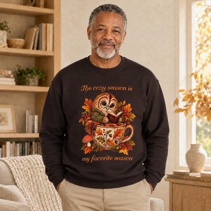 A man wearing a dark brown crewneck sweatshirt with a colorful graphic of an owl in a mug surrounded by autumn leaves, with the text, 'The cozy season is my favorite season'. The man is standing in a cozy, warmly lit living room with bookshelves and a window.
