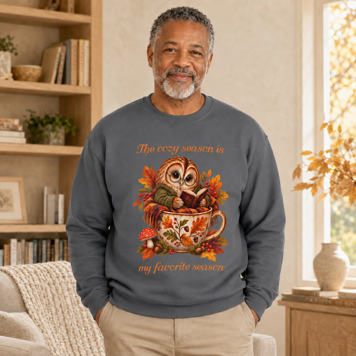 A man wearing a dark gray crewneck sweatshirt with a colorful graphic of an owl reading a book in a teacup surrounded by autumn leaves, with the text, 'The cozy season is my favorite season'. The man is standing in a warm, sunlit living room with bookshelves and a window.