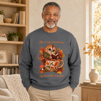 A man wearing a dark gray crewneck sweatshirt with a colorful graphic of an owl reading a book in a teacup surrounded by autumn leaves, with the text, 'The cozy season is my favorite season'. The man is standing in a warm, sunlit living room with bookshelves and a window.