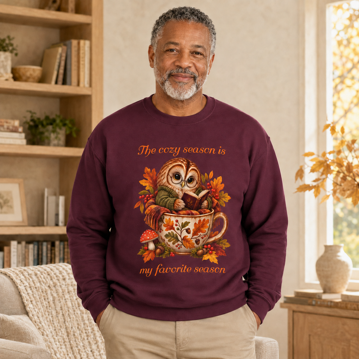 A man wearing a maroon crewneck sweatshirt with a colorful graphic of an owl in a teacup surrounded by autumn leaves, with the text, 'The cozy season is my favorite season'. He is standing in a warmly lit living room with a bookshelf and window in the background.