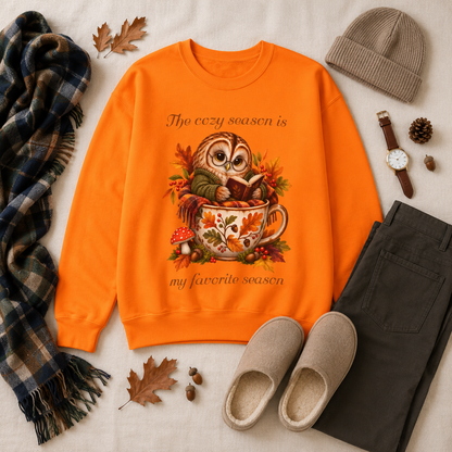 An orange crewneck sweatshirt with an autumn owl graphic and the text, 'The cozy season is my favorite season', styled in a flat lay with a plaid scarf, beanie, watch, pants, slippers, and leaves on a light fabric background.