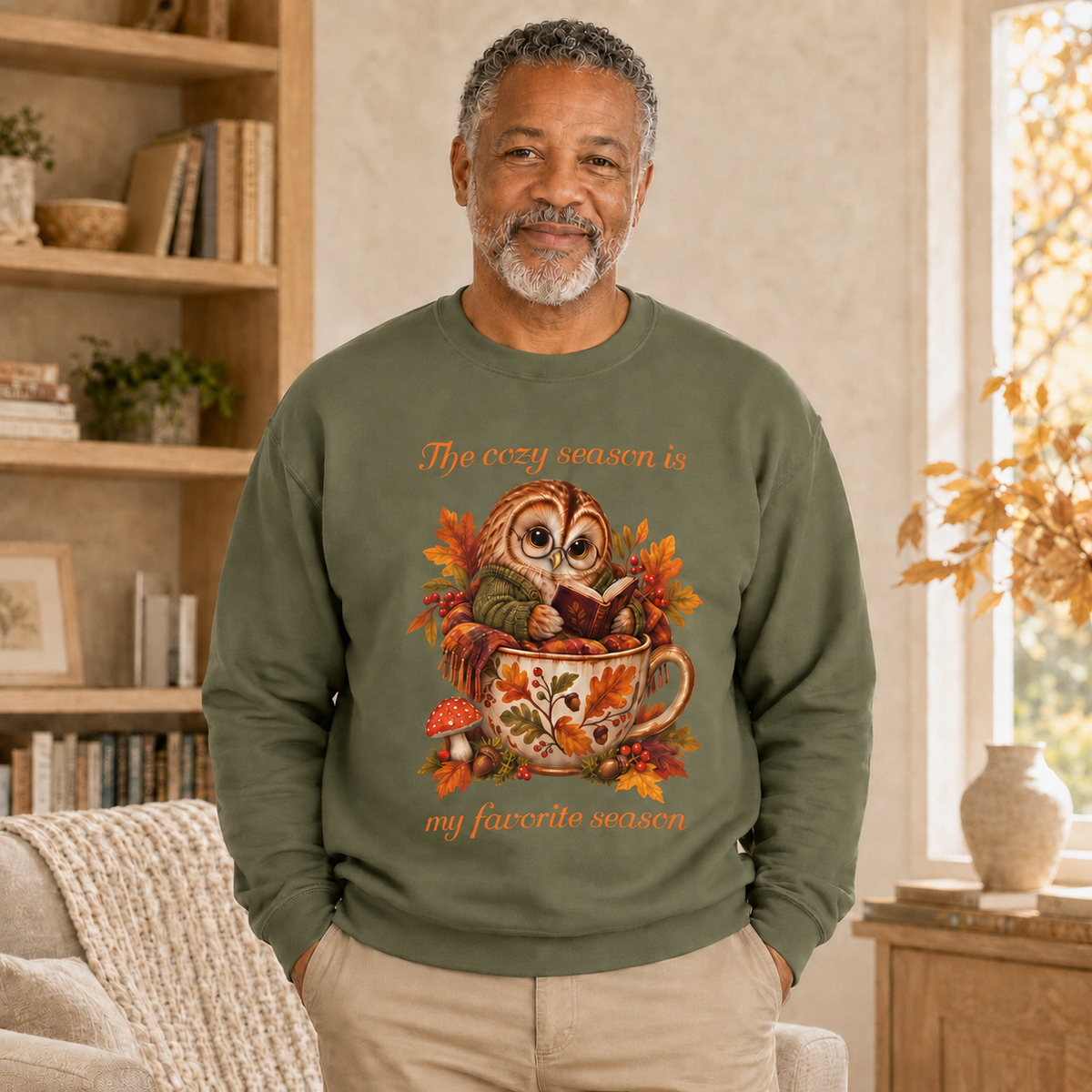 A man wearing an olive green crewneck sweatshirt with a cozy autumn graphic of an owl in a teacup surrounded by leaves. The text on the sweatshirt reads, 'The cozy season is my favorite season'.