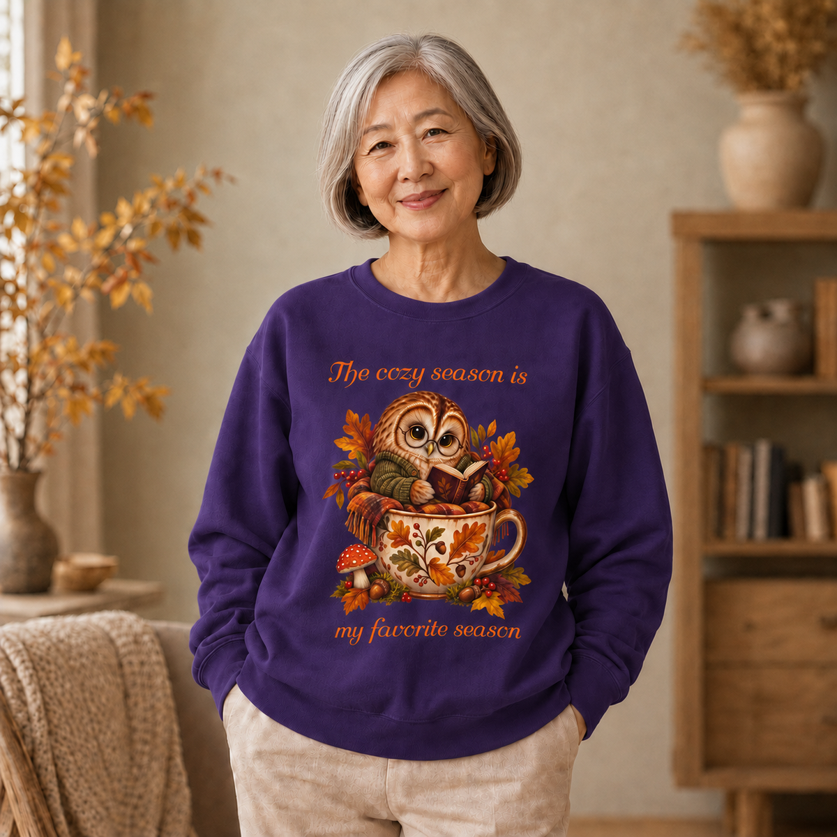 An older woman stands indoors wearing a purple crewneck sweatshirt with a colorful autumn owl graphic and the text, 'The cozy season is my favorite season.' The room has warm neutral tones, a wooden shelf, and dried plants in the background.