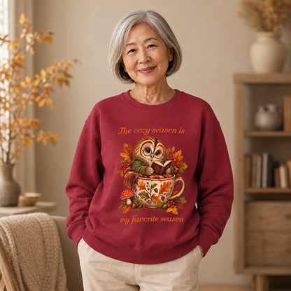An older woman stands indoors wearing a burgundy crewneck sweatshirt with a colorful autumn owl in a teacup graphic and the text 'The cozy season is my favorite season.' The room has warm neutral tones, a bookshelf, and dried floral decor in the background.