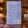 The back cover of Faithful displayed upright on mahogany table with glowing candles, a tea cup, a blanket, and dried lavender flowers around it. The cover is light lavender with black text and muted figures in the background.