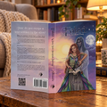 'Fated' displayed open on a wooden table, showing the front cover illustration of Fait, Prometheia, and Broker Rip, alongside the back cover with synopsis text. The setting is a cozy, warmly lit living room with a lamp, potted plants, and coffee mug.