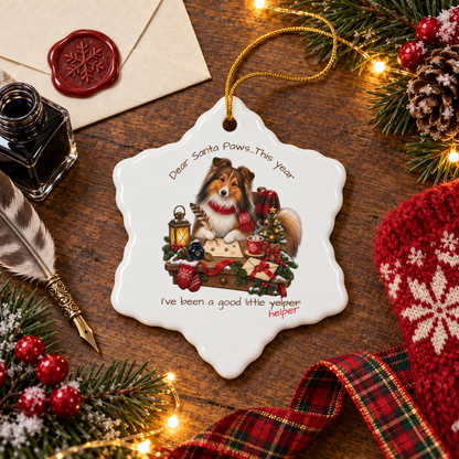 A snowflake-shaped ceramic Christmas ornament featuring a Sheltie dog illustration and the text 'Dear Santa Paws...This year I've been a good little yelper." The last word is crossed out and replaced with 'helper'.