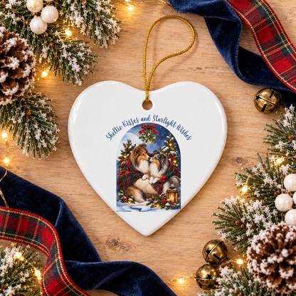 A heart-shaped white ceramic Christmas ornament with a gold hanging loop, featuring a printed illustration of two Shelties in a holiday scene and the text 'Sheltie Kisses and Startlight Wishes.' The ornament is styled on a wooden surface with pine branches, snow-dusted pinecones, gold bells, and red plaid ribbon.