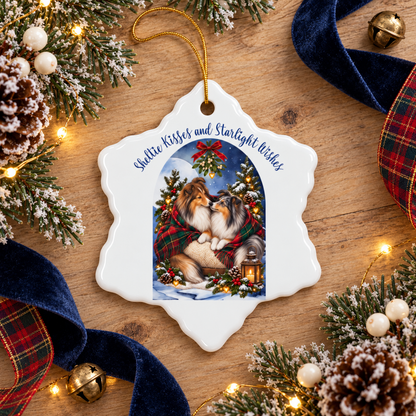 A white snowflake-shaped Christmas ornament with a gold hanging string, featuring an illustration of two Shelties cuddling under mistletoe and the text 'Sheltie Kisses and Starlight Wishes.' The ornament is photographed on a wooden surface surrounded by holiday decorations including pine branches, lights, ribbons, and bells.