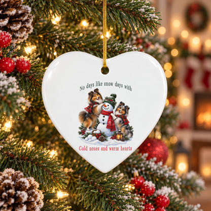 A heart-shaped white ceramic Christmas ornament hanging on a decorated tree. The ornament features a printed illustration of two shelites building a snowman, with the text 'No days like snow days with' above and 'Cold noses and warm hearts' below.