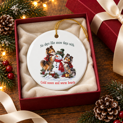 A round white ceramic Christmas ornament with a gold hanging loop, displayed inside a red gift box with a white bow. The ornament features a printed illustration of two shelties building a snowman and the text 'No days like snow days with Cold noses and warm hearts'.
